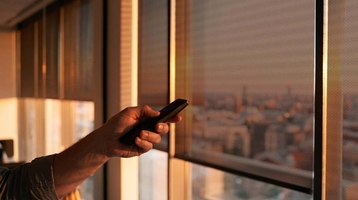Hand with a remote control in front of a roller shade, view of the city at sunset